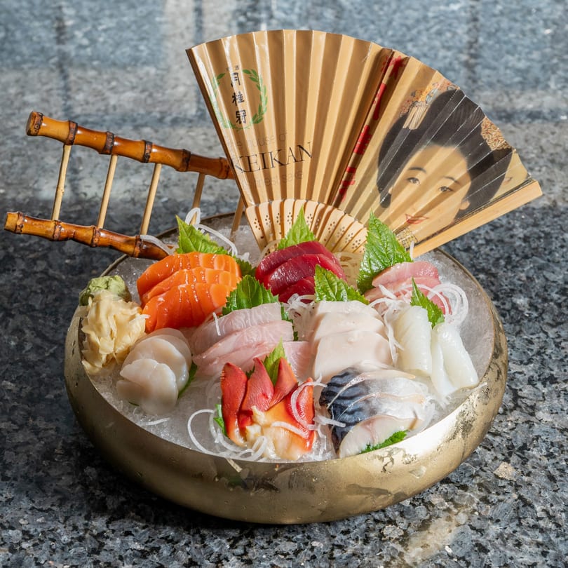 A golden bowl of Kabuki Japanese Cuisine sashimi on ice, with a decorative fan in the background.